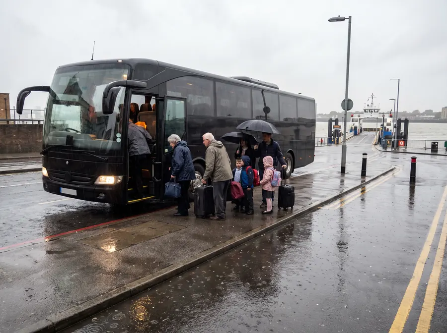 A black executive touring coach parked at the kerb on the approach road to the Woolwich Ferry terminal in steady rain, headlights on and umbrellas up, an extended family of grandparents and children boarding with suitcases, the wide Thames and a ferry beyond.